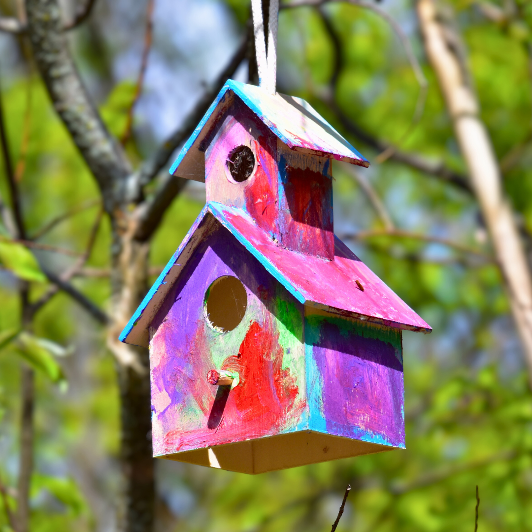 A handmade and painted birdhouse with pinks and blues hangs from a tree.